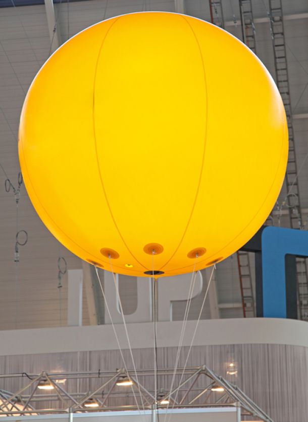 Gelber Riesenballon mit Helium gefüllt, schwebt in einer Messehalle über einem Messestand